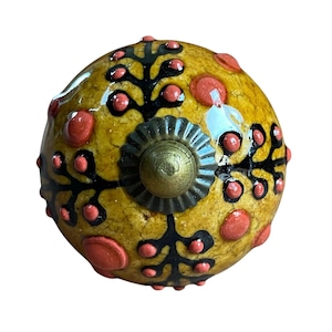 May include: A decorative cabinet knob with a yellow base, decorated with black floral patterns and red accents. The knob has a brass-coloured centre and a textured surface, adding a unique touch to furniture or decor.