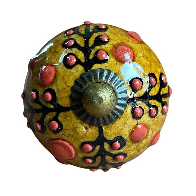 May include: A decorative cabinet knob with a yellow base, decorated with black floral patterns and red accents. The knob has a brass-coloured centre and a textured surface, adding a unique touch to furniture or decor.