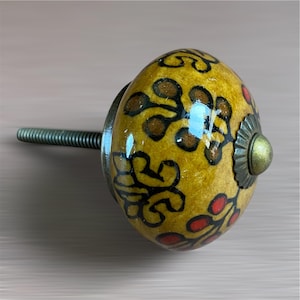 May include: A decorative cabinet knob with a yellow, oval-shaped ceramic design. The knob features a floral pattern in black, brown, and red. It has a metal screw and a decorative metal base with a brass-coloured centre.