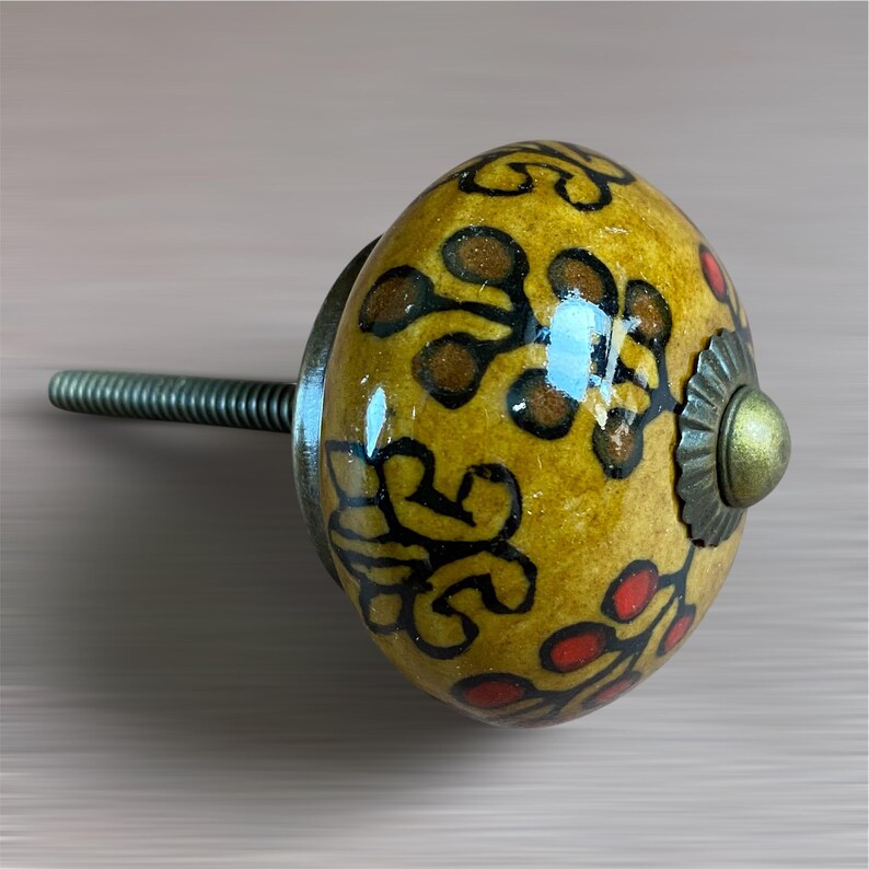 May include: A decorative cabinet knob with a yellow, oval-shaped ceramic design. The knob features a floral pattern in black, brown, and red. It has a metal screw and a decorative metal base with a brass-coloured centre.
