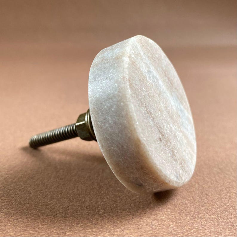 Pode incluir: Um puxador de arm&aacute;rio de m&aacute;rmore de cor clara, em forma oval, com um parafuso de metal roscado e um encaixe de lat&atilde;o. O puxador tem uma superf&iacute;cie lisa e polida, projetado para m&oacute;veis ou arm&aacute;rios.