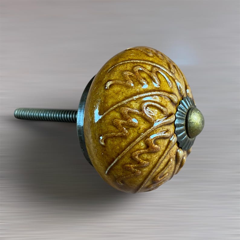 May include: A mustard yellow ceramic cabinet knob with a decorative swirling pattern. The knob has a metal base and screw for fitting. The design is reminiscent of vintage or antique hardware. The knob is suitable for cabinets and drawers.