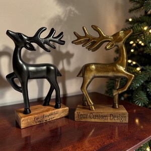 May include: Two standing deer figurines, one matte black and one metallic gold, each featuring large antlers and mounted on a rectangular wooden base. The black base is engraved with "Live Laugh Love" and the gold base with "Your Custom Design."