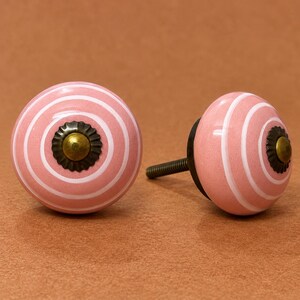 May include: Two round ceramic cabinet knobs featuring a pink surface with concentric white circular stripes. Each knob is centered with a dark bronze-toned metal floral-shaped washer and a small brass-colored dome-shaped screw head in the middle.