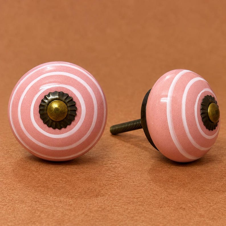 May include: Two round ceramic cabinet knobs featuring a pink surface with concentric white circular stripes. Each knob is centered with a dark bronze-toned metal floral-shaped washer and a small brass-colored dome-shaped screw head in the middle.
