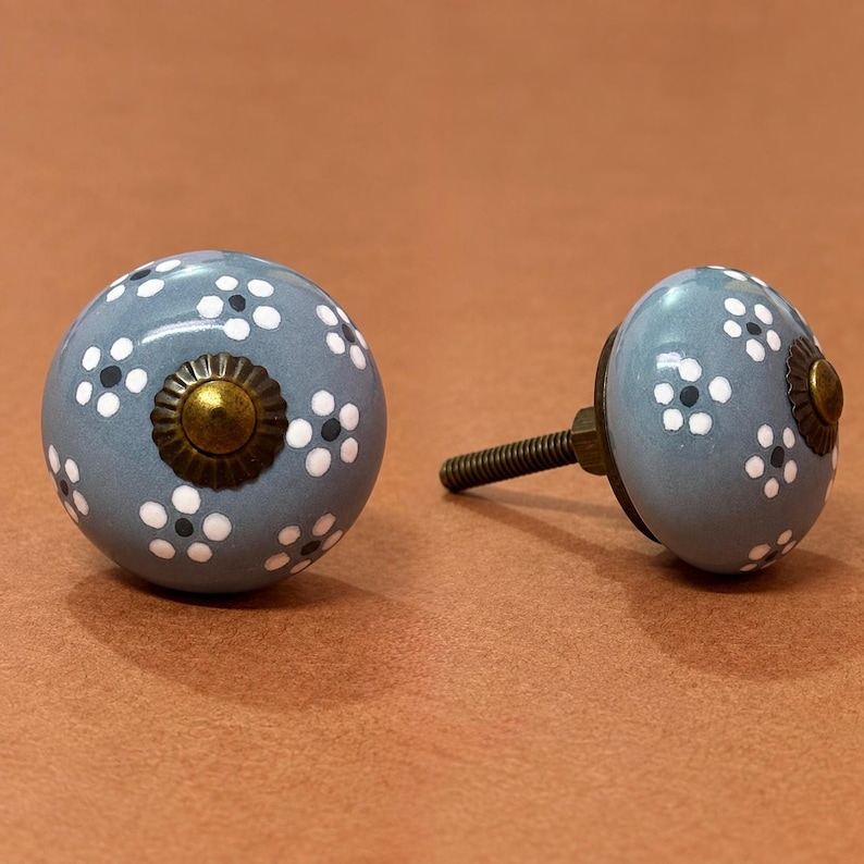 May include: Two round ceramic cabinet knobs in a dusty blue color, each featuring a hand-painted pattern of small white flowers with dark centers. Each knob has a central brass-toned metal cap with a scalloped edge and a threaded metal bolt.