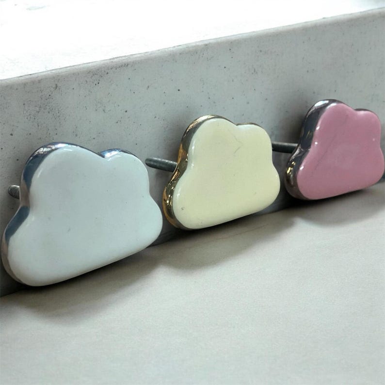 May include: Three cloud-shaped cabinet knobs in a row. The knobs are white, cream, and pink, each with a silver or gold trim. The knobs are made of ceramic or porcelain and are mounted on metal posts.