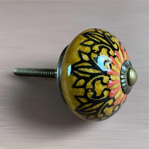 May include: A decorative cabinet knob with a yellow, black, and coral floral design. The ceramic knob has a metal base and a threaded screw for installation. The knob is approximately 5 cm in diameter.