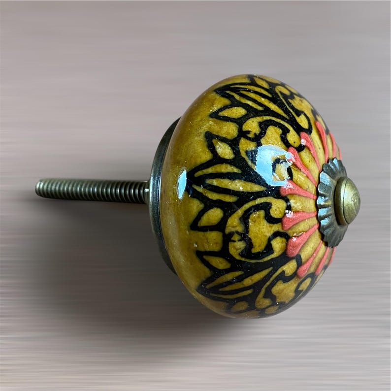 May include: A decorative cabinet knob with a yellow, black, and coral floral design. The ceramic knob has a metal base and a threaded screw for installation. The knob is approximately 5 cm in diameter.