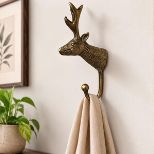 May include: A wall-mounted hook crafted from antique brass-toned metal, featuring a detailed deer head sculpture with prominent antlers. The hook extends downwards from the deer's neck, ending in a rounded tip, currently holding a beige fabric cloth.