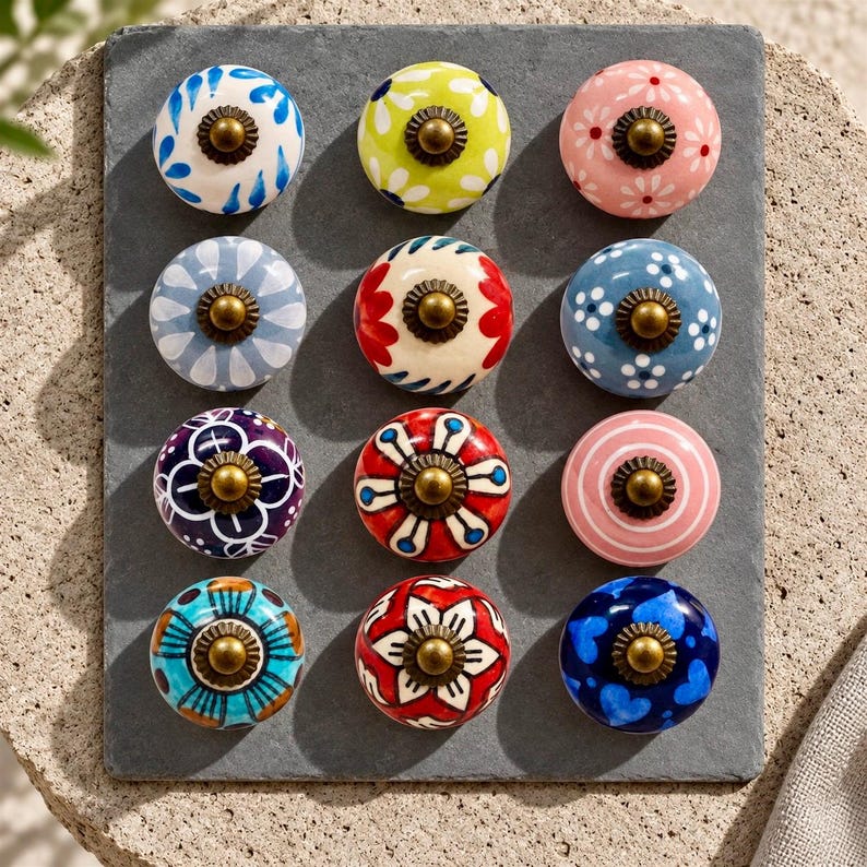 May include: A set of twelve round ceramic cabinet knobs arranged in a grid. Each knob features a unique hand-painted pattern in various colors including blue, red, pink, green, and white, finished with a central antique brass-toned metal screw.