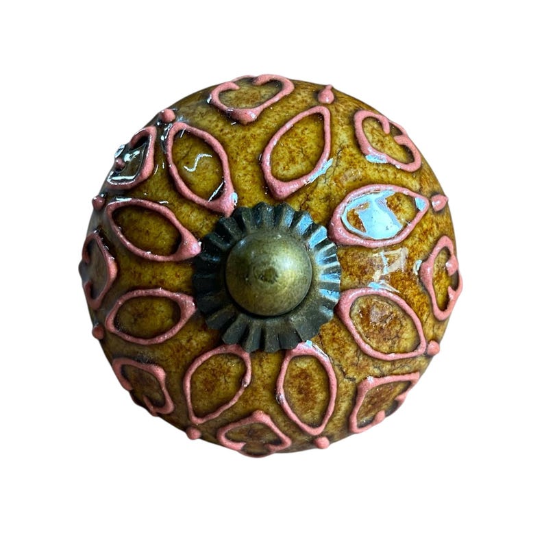 May include: A decorative cabinet knob with a textured, mustard-yellow surface. The knob features a pattern of pink, oval-shaped designs. A bronze-coloured, floral-shaped base and centre detail complete the design. This unique knob is perfect for adding a touch of style to furniture.