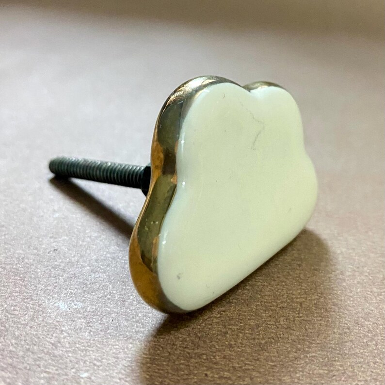 May include: A decorative cabinet knob in a cloud shape. The knob has a creamy white centre with a gold-coloured trim. A threaded screw extends from the back for installation. The knob is designed for furniture and home decor.
