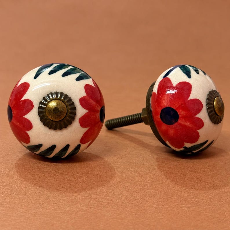 May include: Two round ceramic cabinet knobs featuring a cream-colored base with hand-painted red flower petals and dark green leaf patterns. Each knob has a central antique brass-toned metal cap and a threaded metal bolt extending from the back.