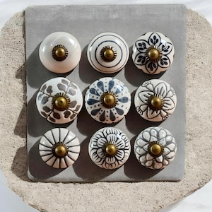 May include: Nine decorative ceramic cabinet knobs in various designs. The knobs are white with patterns in black, gray, and blue. Each knob has a brass-colored center. The knobs are arranged on a gray surface.