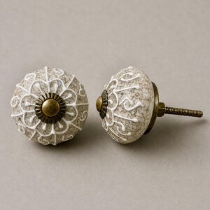 Op de afbeelding: Twee decoratieve kastknoppen. De knoppen zijn rond met een gespikkelde, lichtbruine basis en witte bloemmotieven. Elke knop heeft een messingkleurige metalen basis en een schroefdraad voor installatie.