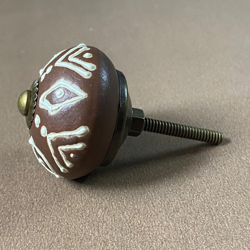 Puede incluir: Un pomo de armario de cer&aacute;mica marr&oacute;n con patrones geom&eacute;tricos blancos. El pomo tiene una base de metal, una arandela de color lat&oacute;n y un perno roscado para la instalaci&oacute;n. El dise&ntilde;o es sim&eacute;trico y decorativo.