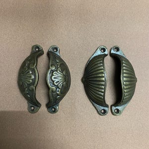 Puede incluir: Cuatro tiradores de armario antiguos de color bronce. Dos tienen un diseño floral y los otros dos tienen un diseño acanalado en forma de abanico. Cada tirador tiene dos orificios para tornillos para el montaje. Los tiradores están dispuestos sobre un fondo neutro.