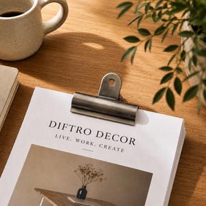 May include: A silver metal binder clip on a stack of white paper with the text "DIFTO DECOR LIVE. WORK. CREATE." A small black vase with dried flowers is visible in the image on the paper. A beige mug is in the background.