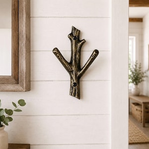May include: A bronze-colored metal wall hook shaped like a tree branch with three prongs. The hook is mounted on a white, horizontal-planked wall. The hook is a decorative and functional item for hanging coats, hats, or other items.