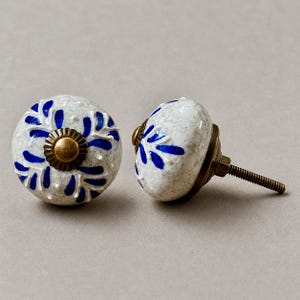 Op de afbeelding: Twee keramische kastknoppen. De knoppen zijn wit met een gespikkeld patroon en blauwe bloemmotieven. Elke knop heeft een messingkleurige basis en een schroefdraad voor installatie. Decoratieve meubelhardware.