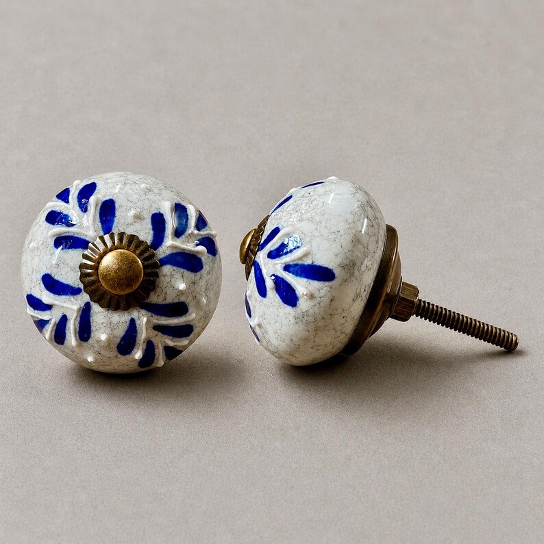 Op de afbeelding: Twee keramische kastknoppen. De knoppen zijn wit met een gespikkeld patroon en blauwe bloemmotieven. Elke knop heeft een messingkleurige basis en een schroefdraad voor installatie. Decoratieve meubelhardware.