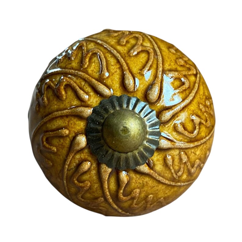 May include: A round, mustard-yellow ceramic cabinet knob with a raised floral design. The knob features a brass-coloured centre and a decorative, dark metal base. The design is detailed and ornate, suitable for furniture or decor.