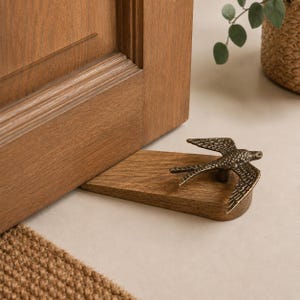 Pode incluir: Um batente de porta em forma de cunha feito de madeira castanha com um ornamento de pássaro em metal cor de bronze no topo. O pássaro é representado com as asas abertas e texturas de penas detalhadas, posicionado para segurar uma porta.