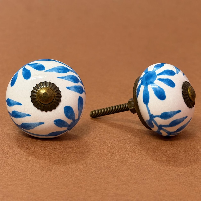 May include: Two round ceramic cabinet knobs featuring a white base with hand-painted blue floral patterns. Each knob has a dark bronze-toned metal center cap and a matching metal screw post extending from the back of one of the knobs.