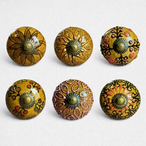 Könnte beinhalten: Sechs dekorative Keramik-Schrankknöpfe in verschiedenen Designs. Die Knöpfe sind hauptsächlich gelb mit aufwendigen Mustern in Braun, Rot und Schwarz. Jeder Knopf hat einen Messingfarbenen Metallsockel.