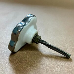 May include: A white ceramic cabinet knob with a silver trim and a threaded metal screw. The knob has a flower shape. The hardware is for furniture and home decor.