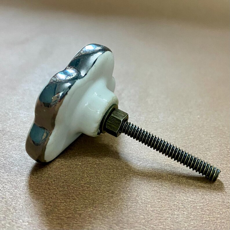 May include: A white ceramic cabinet knob with a silver trim and a threaded metal screw. The knob has a flower shape. The hardware is for furniture and home decor.