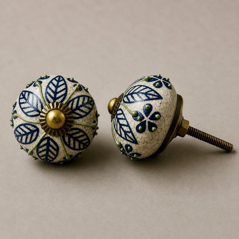 Op de afbeelding: Twee decoratieve keramische kastknoppen. De knoppen zijn gebroken wit met een gespikkelde textuur en hebben een blauw bladeren ontwerp. Elke knop heeft een messingkleurige basis en een schroefdraad voor installatie. Ideaal voor het decoreren van meubels.