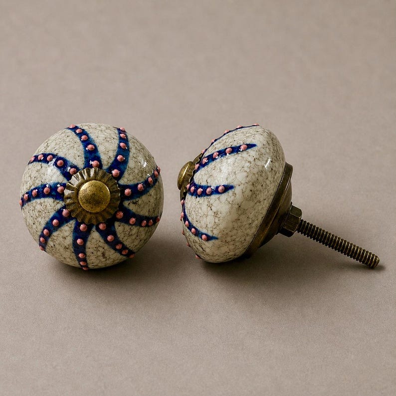 Op de afbeelding: Twee decoratieve kastknoppen. De knoppen zijn rond met een gespikkelde, gebroken witte afwerking. Ze hebben een blauw en roze sterrenpatroon en een messingkleurige basis. De knoppen hebben een schroefdraadbout voor installatie.