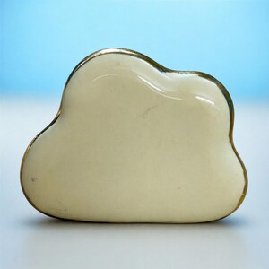 May include: A cream-coloured, cloud-shaped ceramic dish with a gold trim. The dish has a smooth, glossy surface and an organic form. The background is a gradient of blue and white.