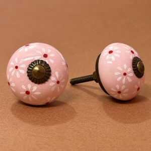 May include: Two round cabinet knobs with a light pink base color, featuring a repeating pattern of white daisy-like flowers with small red centers. Each knob has a central antique brass-toned metal rosette and a threaded metal mounting screw.