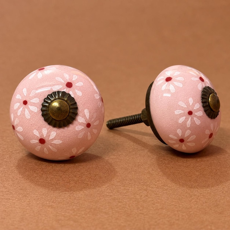 May include: Two round cabinet knobs with a light pink base color, featuring a repeating pattern of white daisy-like flowers with small red centers. Each knob has a central antique brass-toned metal rosette and a threaded metal mounting screw.