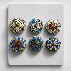 May include: Six decorative ceramic cabinet knobs with floral designs. Each knob features a unique pattern in vibrant colors like blue, orange, yellow, and red, with a brass-colored center. The knobs are arranged on a white surface.