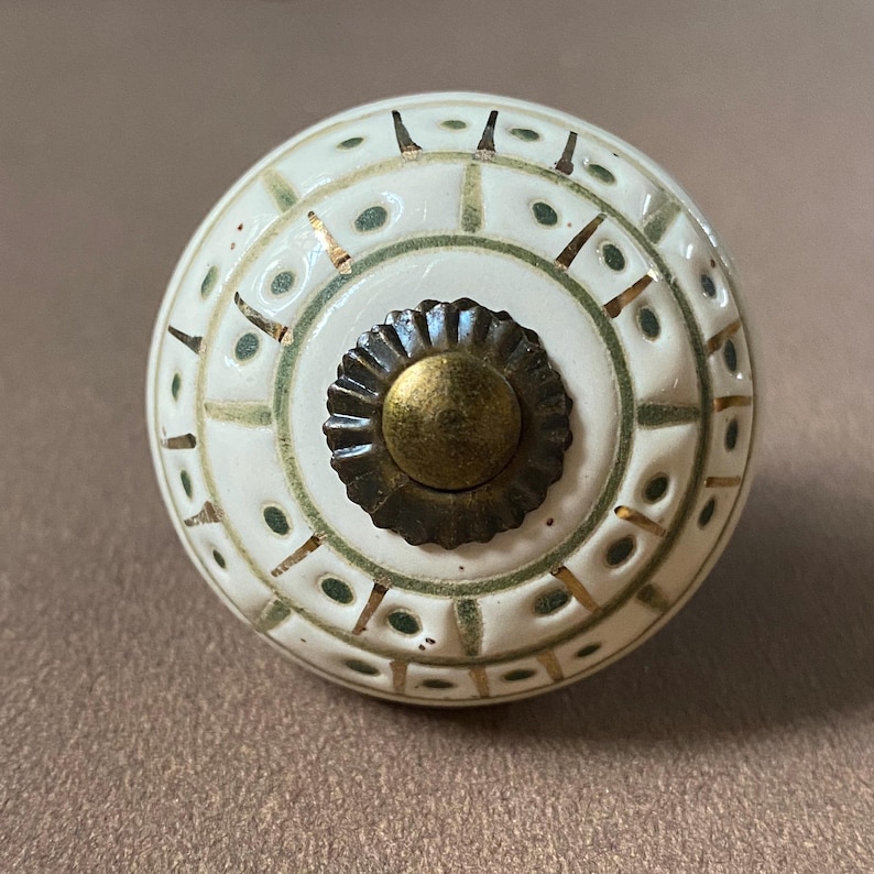 Puede incluir: Pomo de armario de cer&aacute;mica blanca con un dise&ntilde;o decorativo en verde y dorado. El pomo tiene un centro color lat&oacute;n y mide aproximadamente 4 cm de di&aacute;metro. El dise&ntilde;o presenta formas geom&eacute;tricas y puntos.