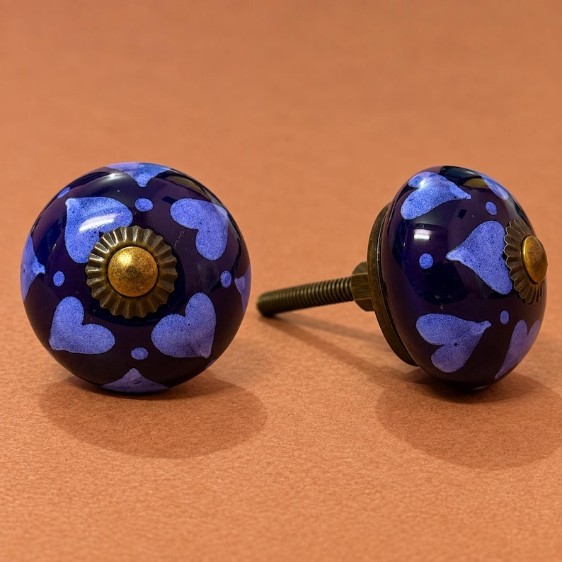 May include: Two round ceramic cabinet knobs featuring a deep navy blue base color with a repeating pattern of light periwinkle blue hearts and small dots. Each knob has a central brass-toned metal rosette and a threaded metal mounting screw.