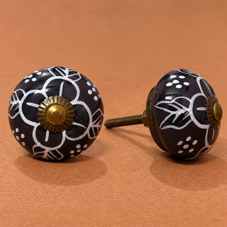 May include: Two round cabinet knobs with a dark black finish and hand-painted white floral patterns. Each knob features a central brass-toned metal cap and a matching brass-toned screw post extending from the back for mounting purposes.