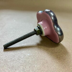 May include: A decorative cabinet knob with a pink, heart-shaped ceramic face and a silver-coloured trim. The knob is attached to a threaded metal rod with a nut and washer. The hardware is designed for furniture and home decor.