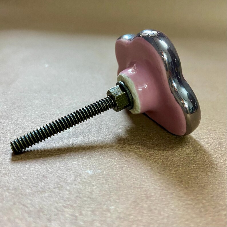 May include: A decorative cabinet knob with a pink, heart-shaped ceramic face and a silver-coloured trim. The knob is attached to a threaded metal rod with a nut and washer. The hardware is designed for furniture and home decor.