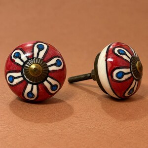 May include: Two round ceramic cabinet knobs featuring a hand-painted floral pattern with red petals, white outlines, and blue centers. Each knob has a central brass-toned metal cap and a threaded metal bolt extending from the back for installation.