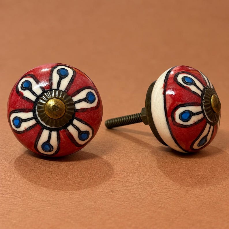 May include: Two round ceramic cabinet knobs featuring a hand-painted floral pattern with red petals, white outlines, and blue centers. Each knob has a central brass-toned metal cap and a threaded metal bolt extending from the back for installation.