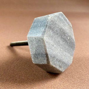 Pode incluir: Um puxador de arm&aacute;rio cinza octogonal com um parafuso de metal roscado. O puxador tem uma apar&ecirc;ncia marmorizada e &eacute; feito de pedra. O hardware foi projetado para uso em gavetas, arm&aacute;rios ou outros m&oacute;veis. O puxador &eacute; mostrado em um fundo marrom.