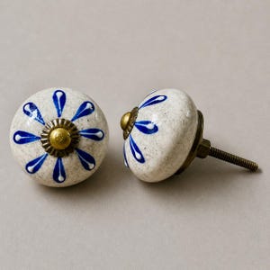 Op de afbeelding: Twee keramische kastknoppen met een witte gespikkelde afwerking en blauw bloemmotief. Elke knop heeft een messingkleurige basis en schroef. De knoppen zijn rond en hebben een decoratieve, handgemaakte uitstraling, geschikt voor meubels of woondecoratie.