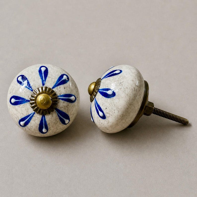 Op de afbeelding: Twee keramische kastknoppen met een witte gespikkelde afwerking en blauw bloemmotief. Elke knop heeft een messingkleurige basis en schroef. De knoppen zijn rond en hebben een decoratieve, handgemaakte uitstraling, geschikt voor meubels of woondecoratie.