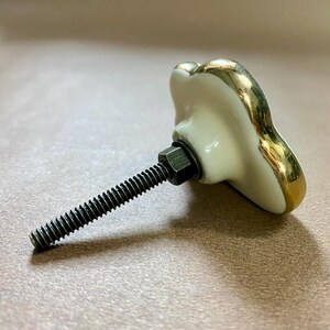 May include: A decorative cabinet knob with a white porcelain finish and gold trim. The knob has a unique shape and is attached to a threaded rod with a hex nut. The hardware is designed for easy installation.
