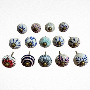 Puede incluir: Una colección de quince pomos de cerámica decorativos para armarios. Cada pomo es redondo con un vástago de latón y presenta un diseño único pintado a mano en varios colores, incluyendo azul, rosa, blanco y rojo. Los pomos están dispuestos sobre un fondo blanco.